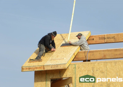 Two men attach a roof panel to a center beam, purlin and exterior wall