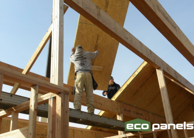 Two men guide a roof panel into place, spanning from ridge beam to exterior wall.